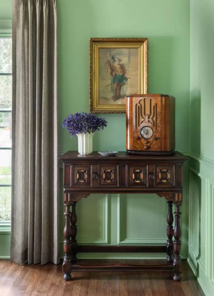 Chiếc radio cổ đặt trên bàn console gỗ trở thành điểm nhấn của góc phòng do Emily Spanos thiết kế. Tường xanh nhạt, rèm vải màu xám, tranh khung mạ và đồ gỗ tối màu tạo sắc thái hoài cổ, trong khi thiết bị nghe đài cơ học thay cho màn hình giúp không gian giữ tinh thần analog, giảm sự hiện diện của công nghệ số. Ảnh: Claudia Casbarian.