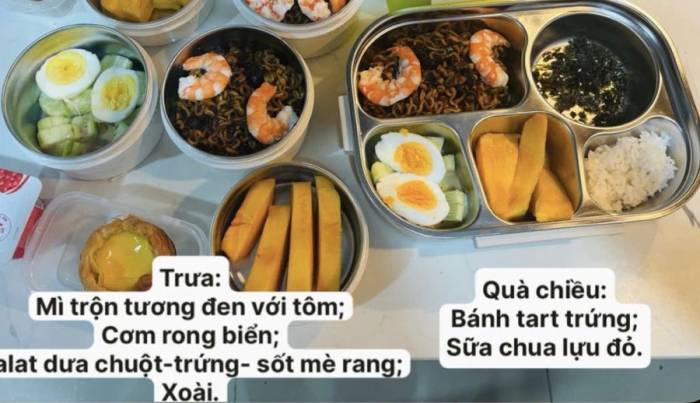 Món mì trộn tương đen, điểm xuyết thêm những con tôm luộc bóc vỏ tươi ngon. Đi kèm với đó là khay cơm trắng ăn cùng rong biển sấy vụn.Để cân bằng dinh dưỡng, bữa ăn được bổ sung thêm món salad dưa chuột tươi mát thanh nhẹ ăn kèm trứng luộc bổ đôi cùng sốt mè rang. Cuối cùng, món tráng miệng là những miếng xoài chín vàng.Bữa quà chiều, phần ăn nhẹ gồm có những chiếc bánh tart trứng vỏ giòn rụm với phần nhân vàng óng béo ngậy, kết hợp cùng các hộp sữa chua vị lựu đỏ đóng gói sẵn vừa thơm ngon vừa tốt cho sức khỏe.Ảnh: Trang Dang